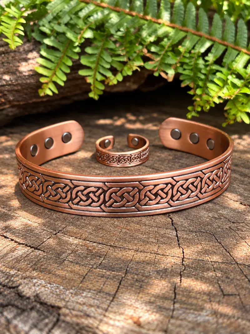 A copper-toned cuff bracelet and a matching ring with an engraved Celtic knot pattern with magnets, displayed on a weathered wood log with green fern leaves in the background.