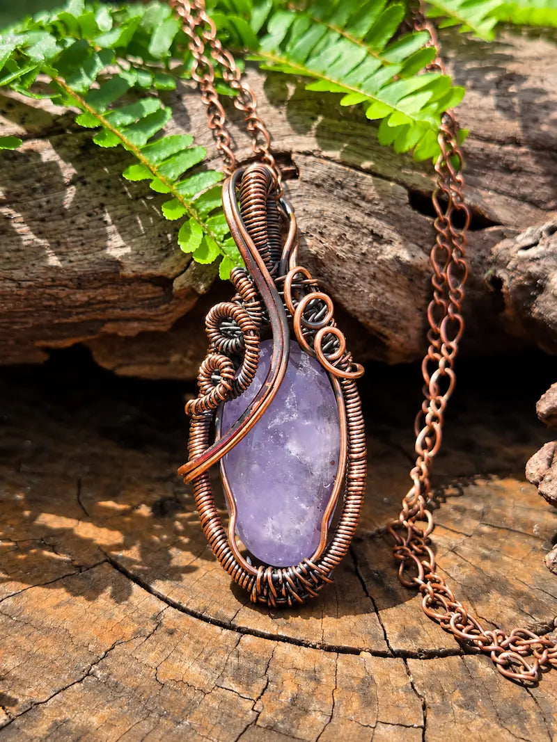 Copper pendant necklace with a amethyst stone, resting on a natural wooden surface with green ferns in the background