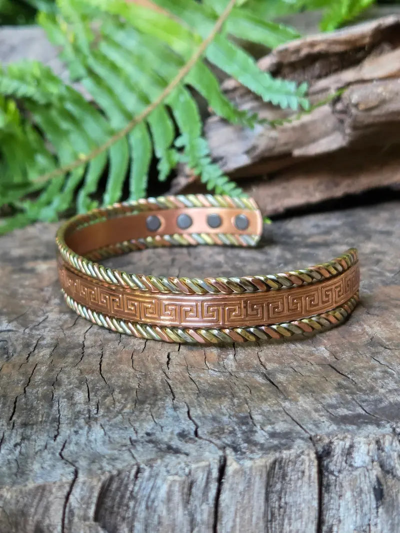 A copper bracelet with a Tricolour greek key design, resting on a natural wooden surface with green ferns in the background.