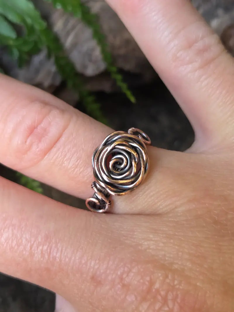 A close-up of a hand wearing a handmade copper ring in a rose gold and dark metallic finish, with a blurred background of green leaves and rocks.