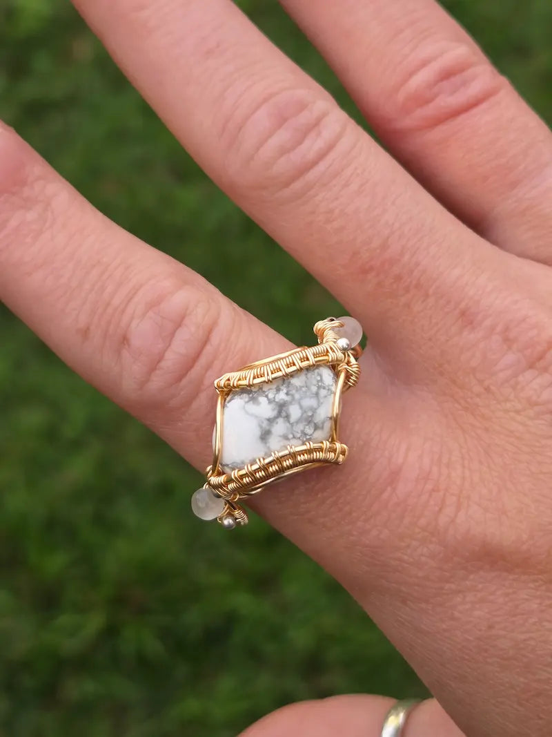 A gold copper wire-wrapped ring featuring a square white and gray marbled stone, worn on a person's finger with a blurred green outdoor background.