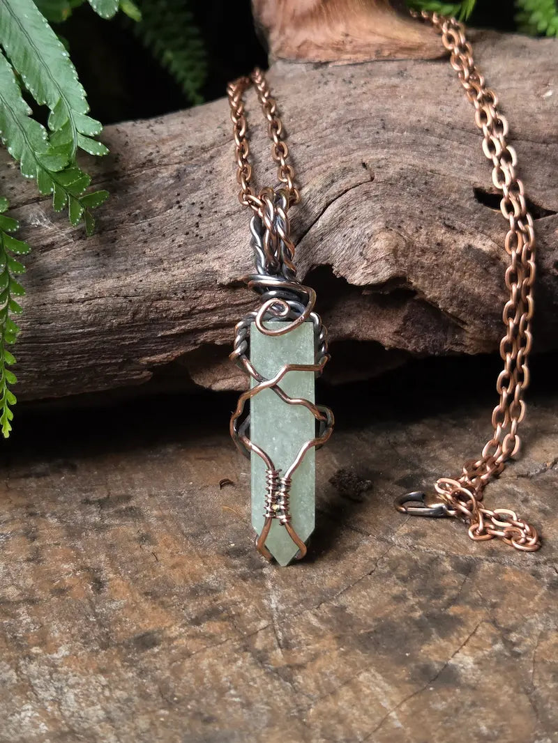 A handmade wire-wrapped pendant necklace featuring a light green crystal point on a copper chain, styled on a rustic piece of wood with green fern leaves.