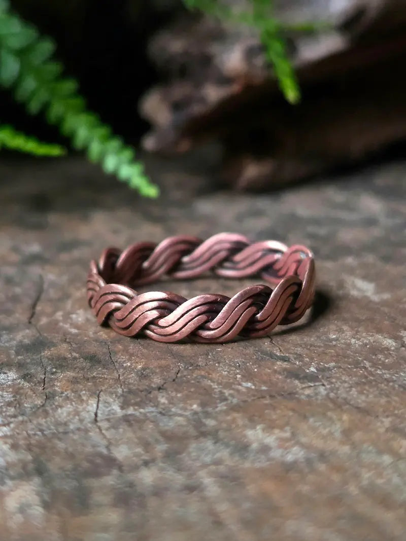 A copper-toned braided ring with a textured, woven pattern, resting on a rustic stone surface with blurred green foliage in the background.