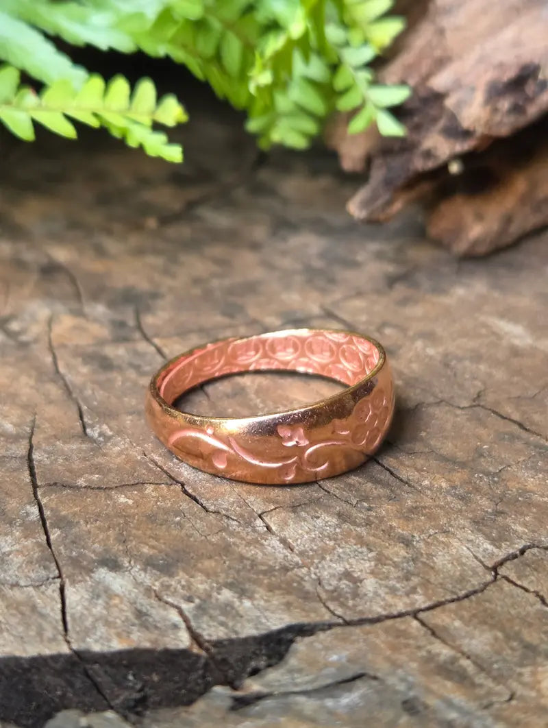 A pure copper ring with a floral pattern, resting on a textured wooden surface with green leaves in the background.