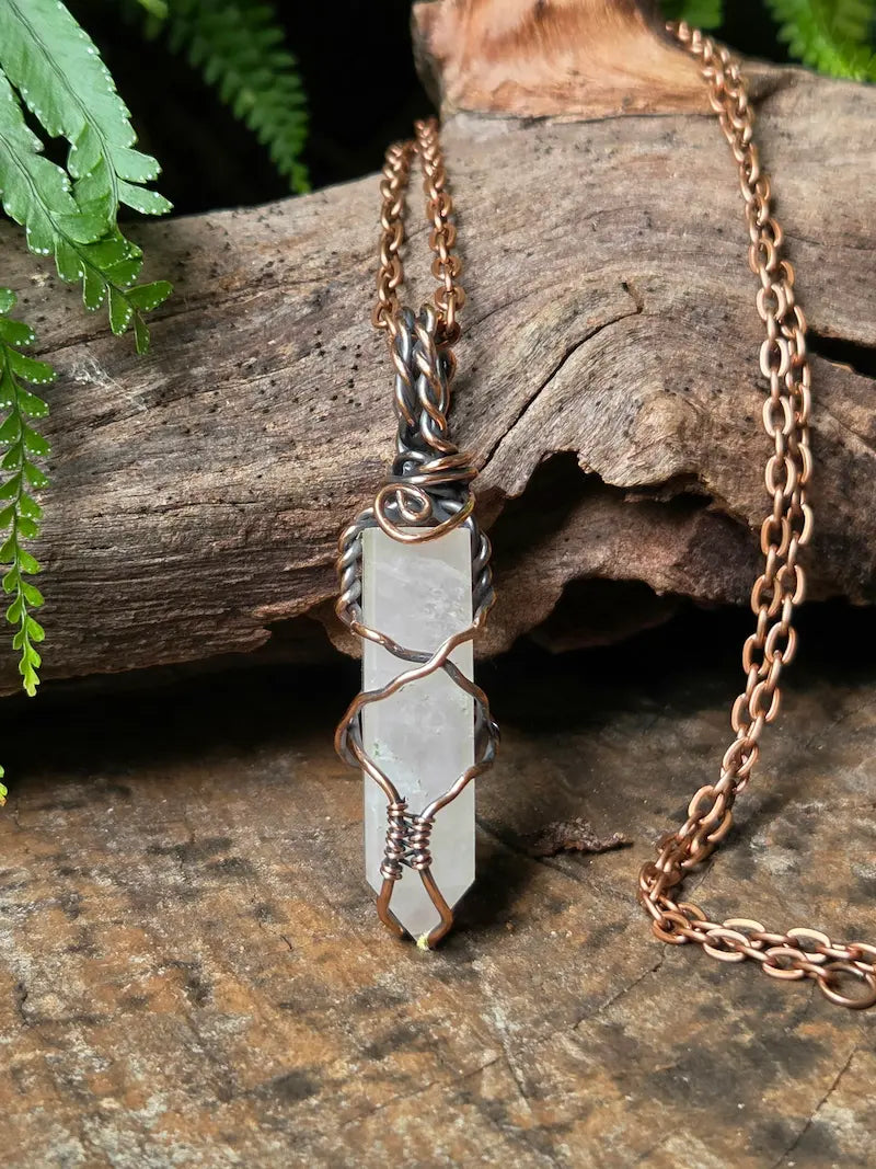 A handmade wire-wrapped pendant necklace featuring a pink crystal point, suspended from a copper chain and displayed on a rustic wood log with green ferns in the background.