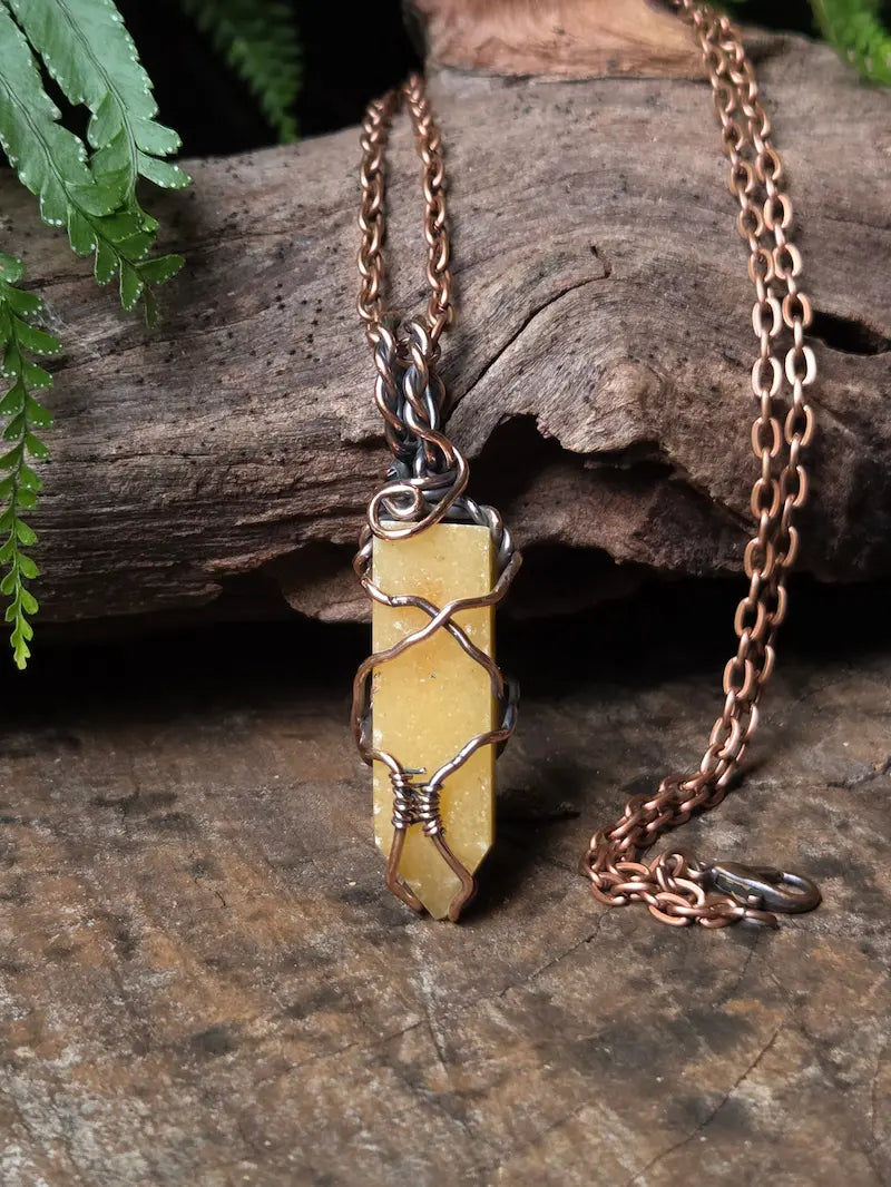 A handmade pendant necklace featuring a yellow stone wrapped in copper wire, hanging from a copper chain on a rustic wood background.