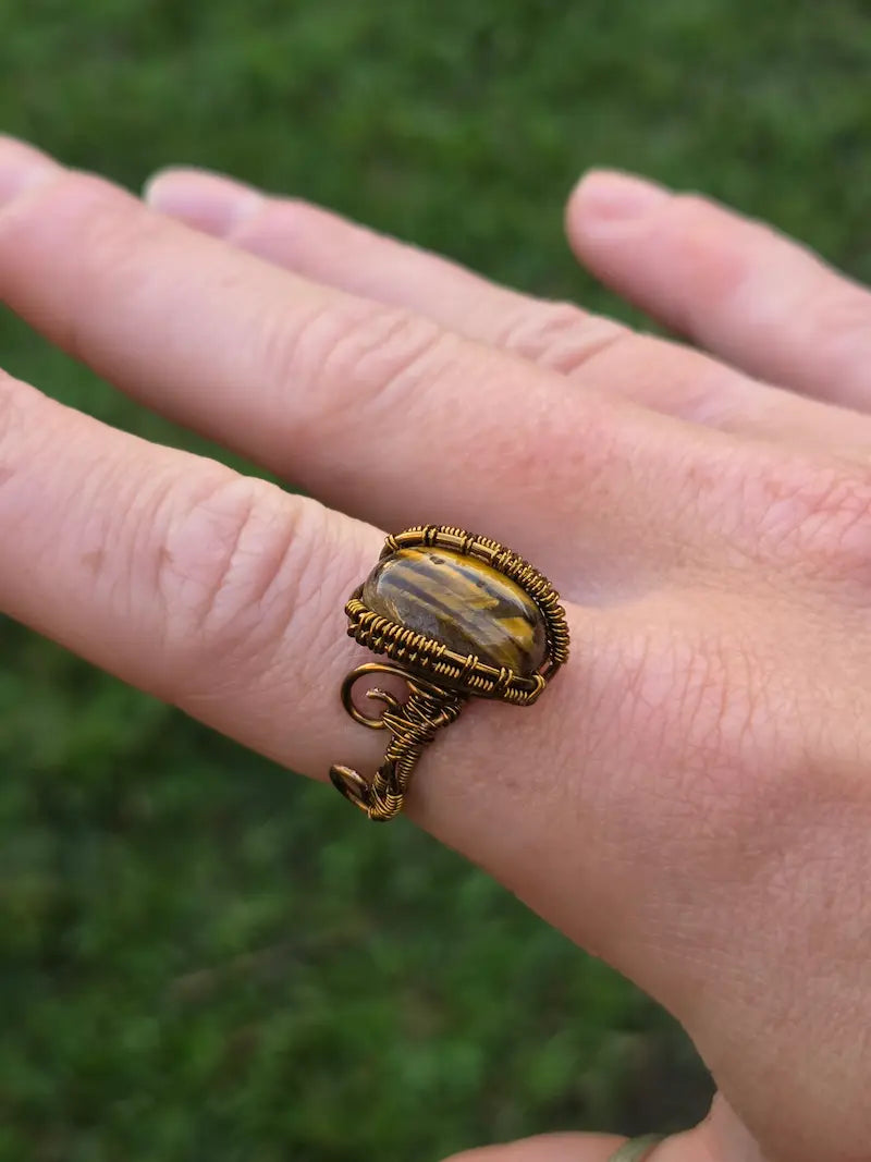 A close-up of a hand wearing a handmade wrap ring with a tiger's eye stone and textured bronze metal, set against a blurred green outdoor background.