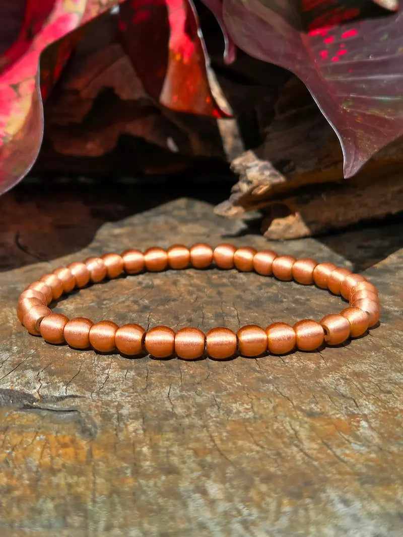 A beaded stretch bracelet made of smooth, rounded copper-colored beads, displayed on a textured wooden surface with red and brown leaves in the background.