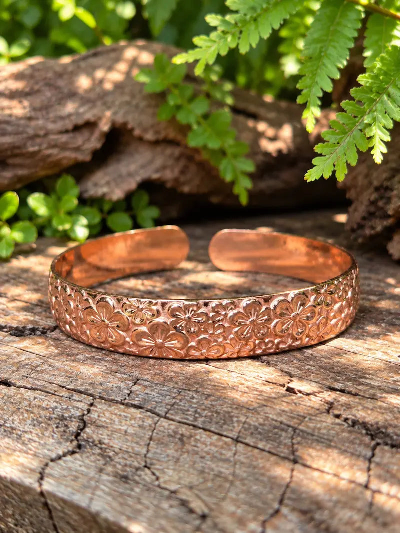 A copper bracelet with a flower design, resting on a natural wooden surface with green leaves in the background