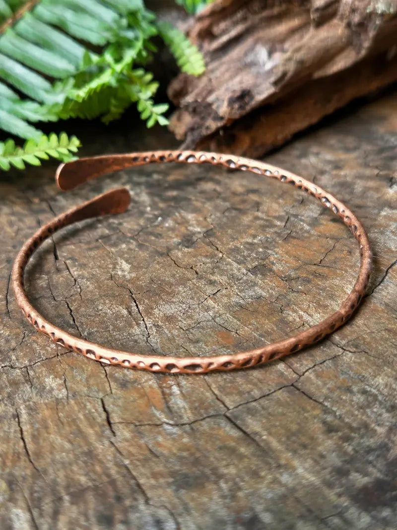 A hammered copper open cuff bracelet resting on a textured wooden surface with green fern leaves and a piece of driftwood in the background.