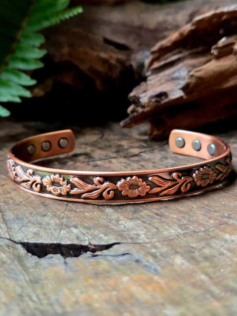 Copper bracelet with flower designs on a natural wooden surface with green ferns in the background