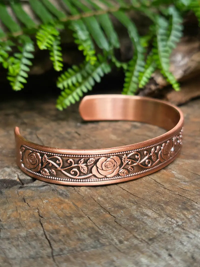Copper bracelet with a elegant floral rose design on a natural wooden surface with green leaves in the background
