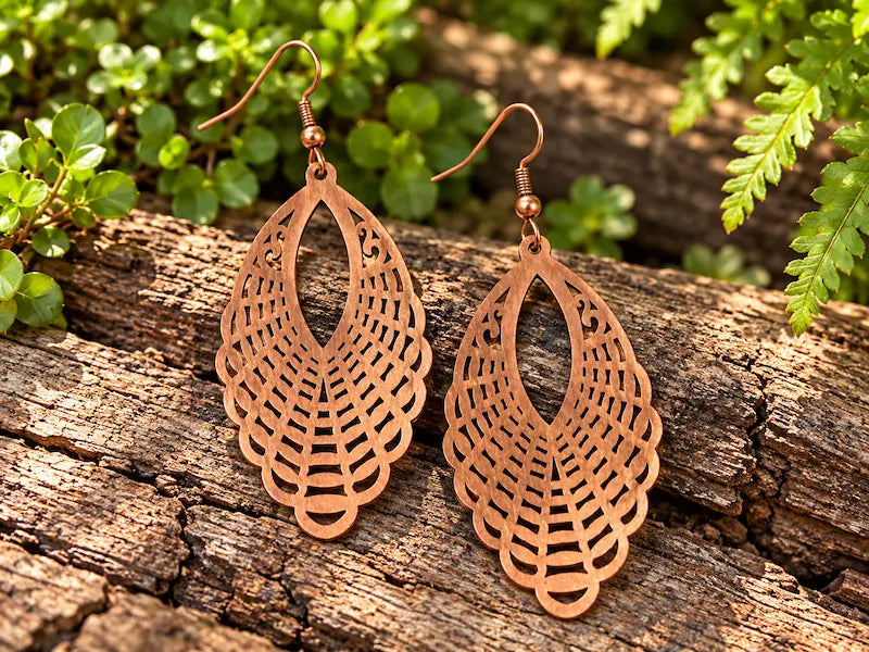 Copper Earrings with a filigree feather design, resting on a natural wooden surface with green leaves in the background