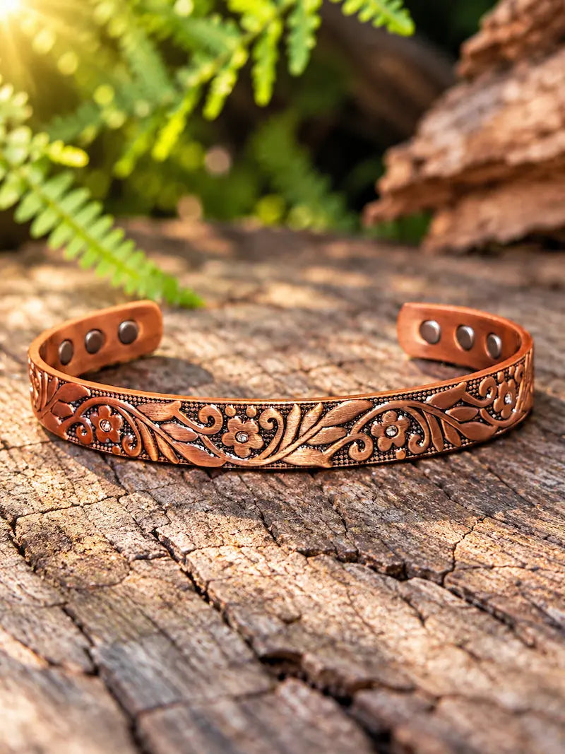 Copper bracelet with a floral pattern, resting on a natural wooden surface with green ferns in the background