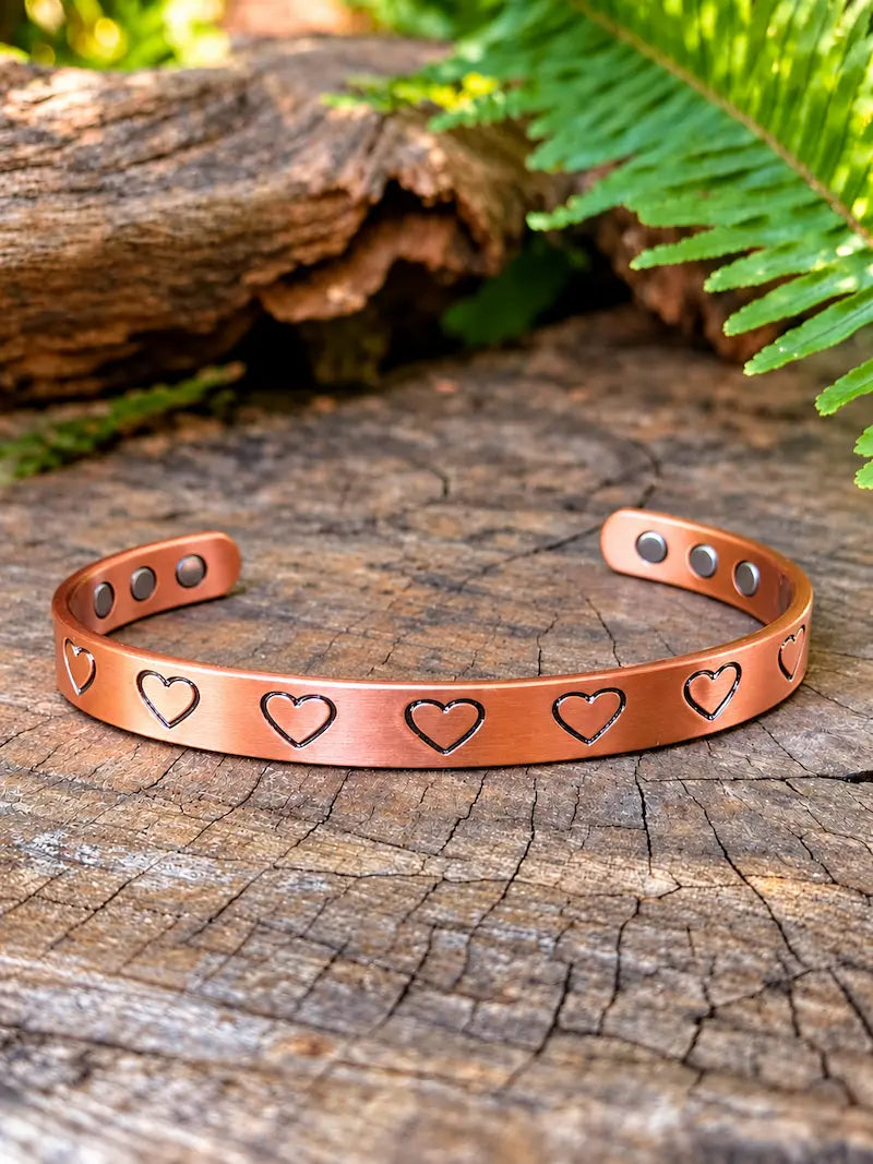 Copper bracelet with a heart design, resting on a natural wooden surface with green ferns in the background