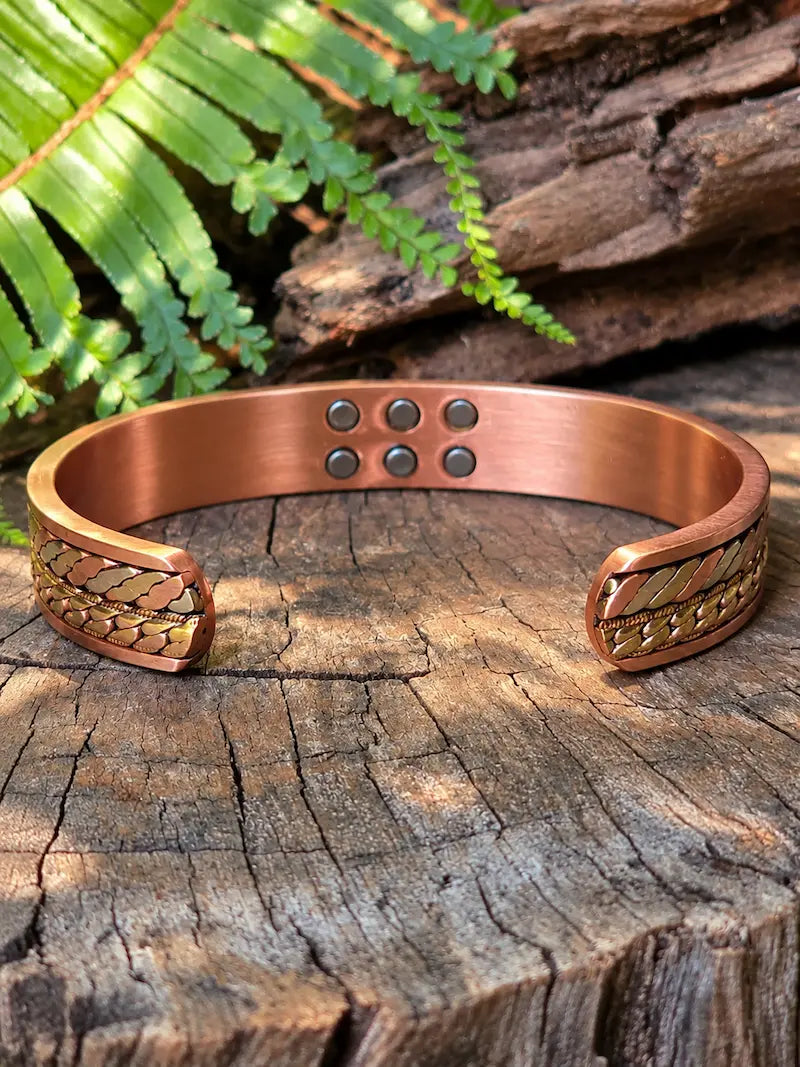 Copper bracelet with a tricolour twisted Chain-Link design, resting on a natural wooden surface with green ferns in the background.
