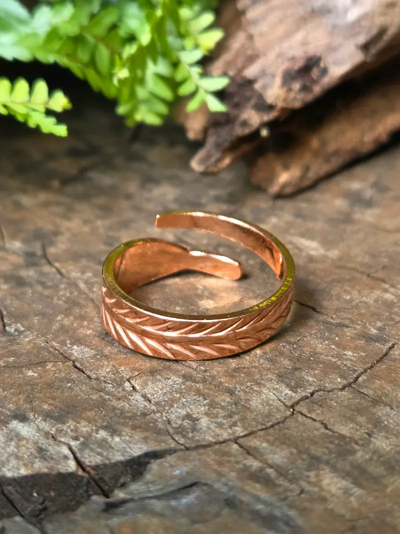 A copper ring with textured leaf-like detailing, resting on a rustic wood surface with blurred green leaves and bark in the background.