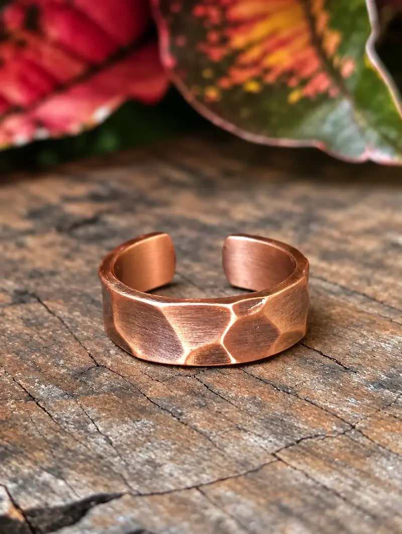 A copper ring with a hammered design, resting on a natural wooden surface with red leaves in the background