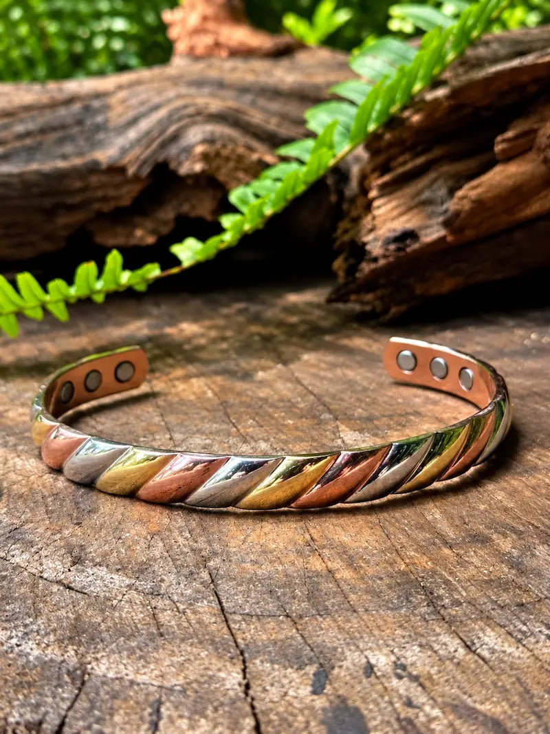 A twisted metallic cuff bracelet with a multi-toned finish of gold, silver, and copper, resting on a rustic wooden log with green ferns in the background.