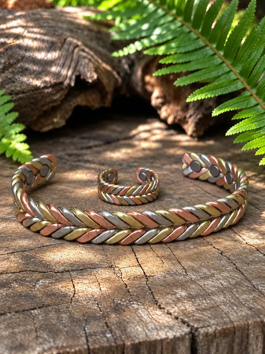A braided cuff bracelet and matching ring with a multi-metal twisted design in copper, gold, and silver, displayed on a rustic wooden log with green ferns in the background.