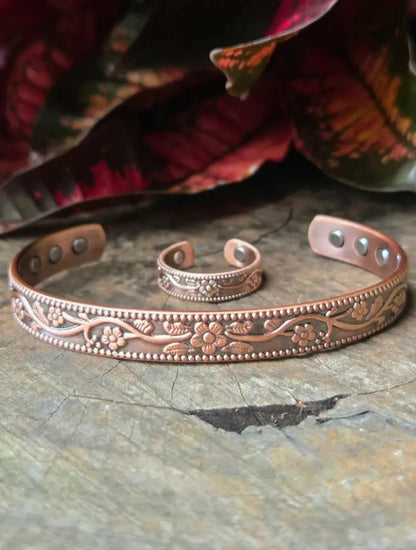 opper cuff bracelet and ring with embossed floral and scroll patterns, displayed on a rustic wooden surface with blurred red and green leaves in the background.