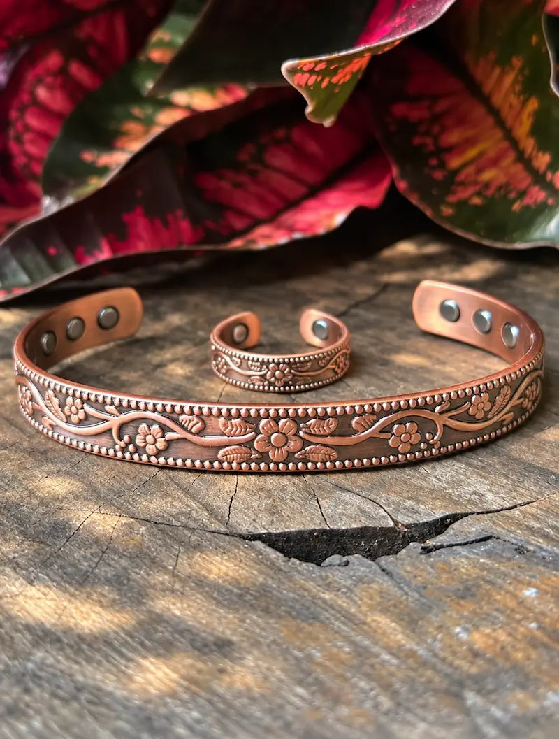 A copper-toned engraved cuff bracelet with a matching ring, displayed on a rustic wooden surface with blurred red and green leaves in the background.