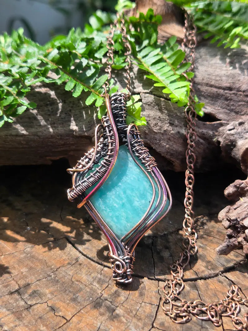 Copper pendant with a Amazonite stone, resting on a natural wooden surface with green ferns in the background 
