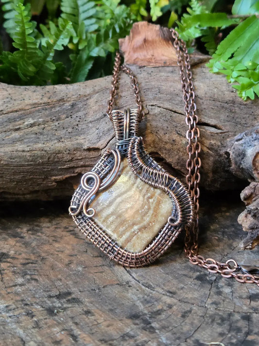 Copper wire-wrapped pendant with a stone on a wooden surface with ferns in the background