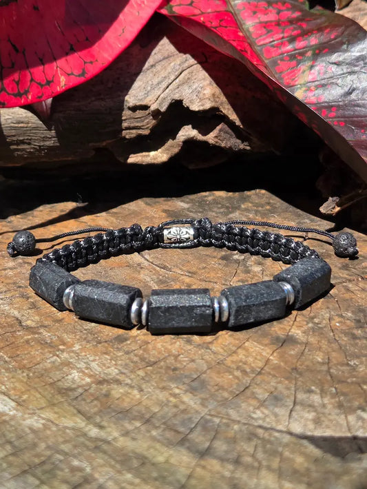 A black beaded bracelet with black Tourmaline beads, silver spacer rings, and a black cord slider clasp, displayed on a rustic wooden surface with red leaves in the background.
