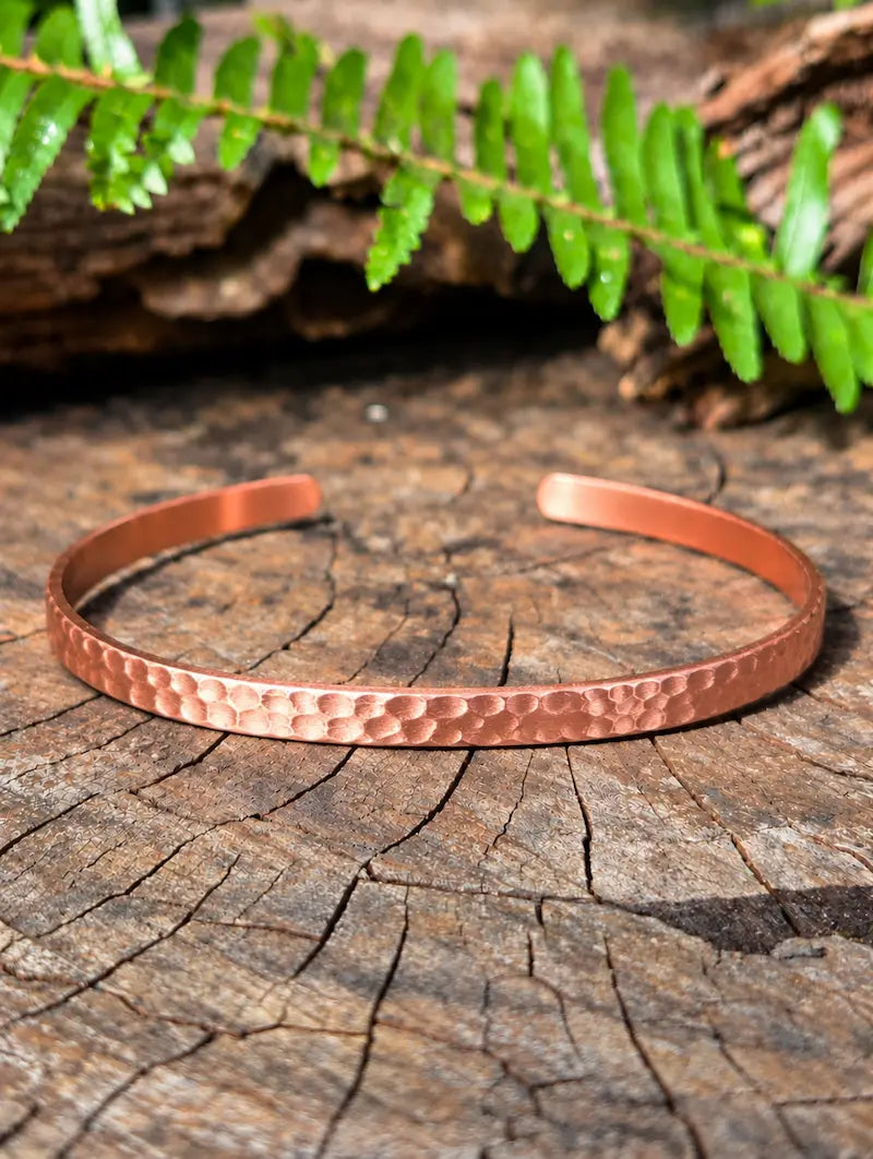 Copper bracelet with a hammered design, resting on a natural wooden surface with green ferns in the background 