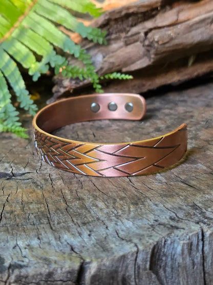 A copper bracelet with a sunburst geometric design, resting on a natural wooden surface with green ferns in the background.
