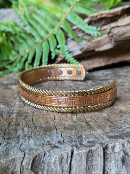 A copper bracelet with a Tricolour greek key design,  resting on a natural wooden surface with green ferns in the background.