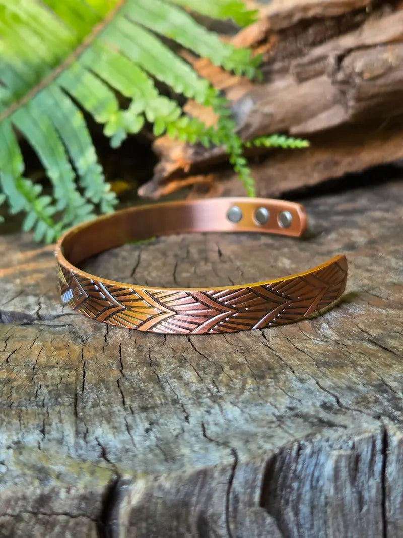 A copper bracelet with a geometric design, resting on a natural wooden surface with green ferns in the background 