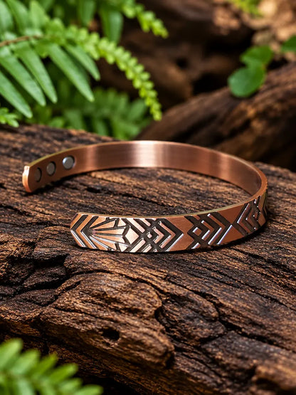 Copper Bracelet with a geometric pattern resting on a natural wooden surface with green ferns in the background 