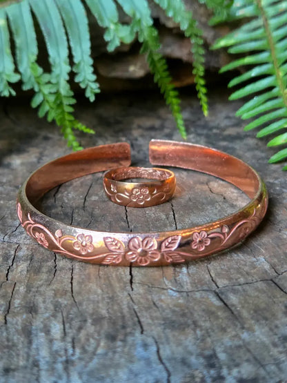 A copper open cuff bracelet with engraved floral patterns, displayed on a weathered wood surface with a matching ring and green fern leaves in the background.