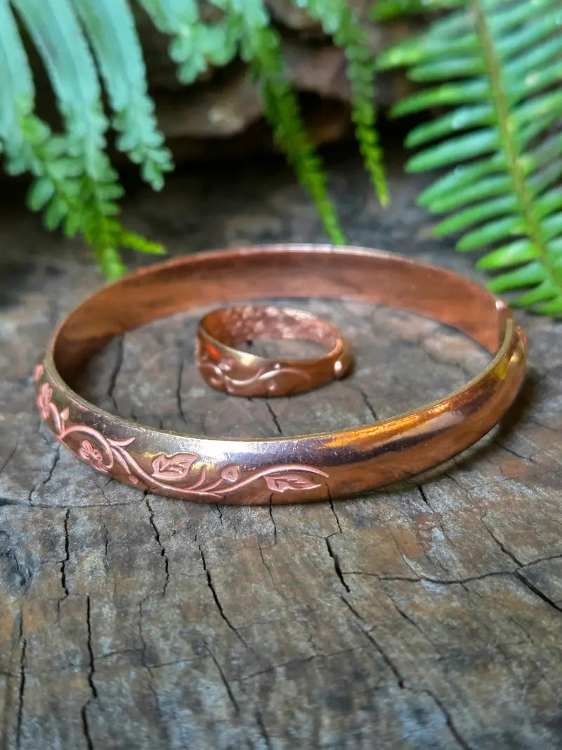 A copper bracelet with engraved floral patterns, displayed on a rustic wooden surface with a matching copper ring behind it and green fern leaves in the background.
