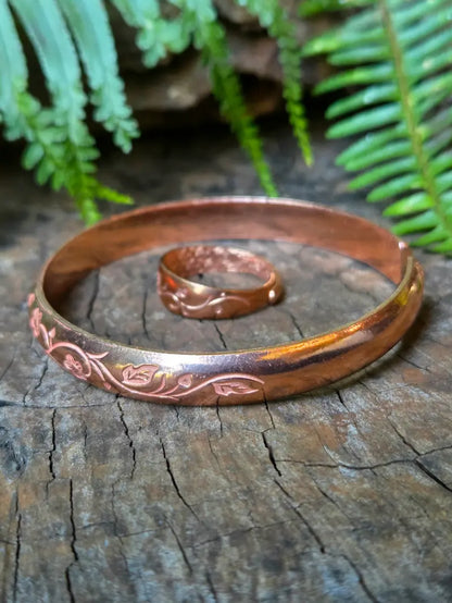 A copper bracelet with engraved floral patterns, displayed on a rustic wooden surface with a matching copper ring behind it and green fern leaves in the background.