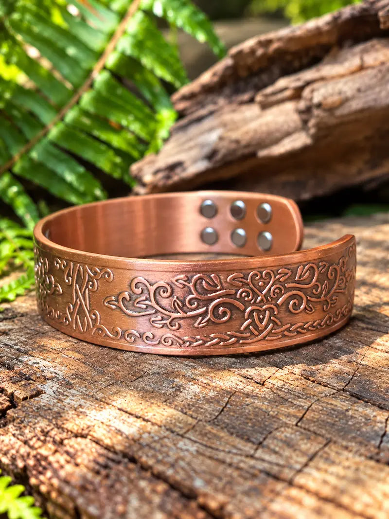 Copper bracelet with a vine scrollwork design, resting on a natural wooden surface with green ferns the background 
