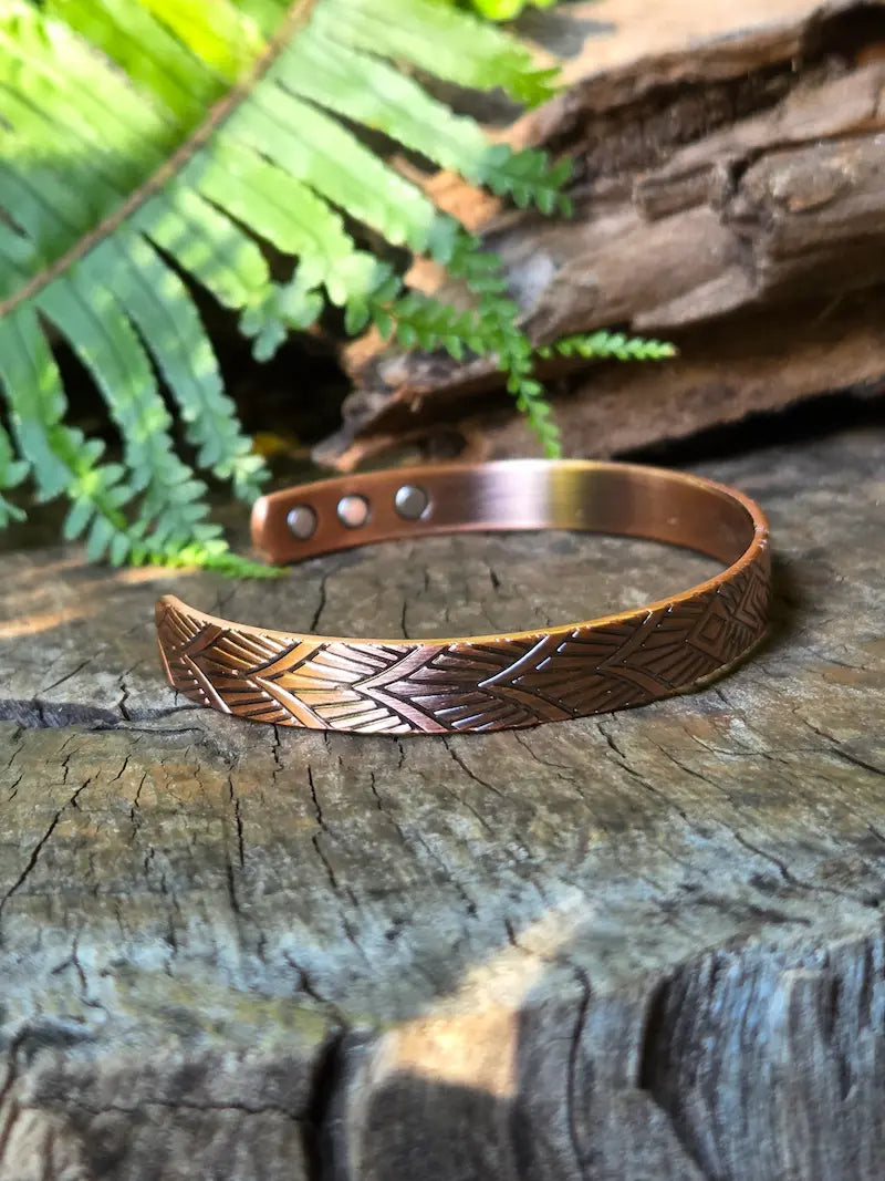 A copper bracelet with a geometric design, resting on a natural wooden surface with green ferns in the background 