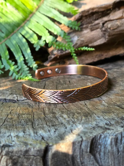 A copper bracelet with a geometric design, resting on a natural wooden surface with green ferns in the background 