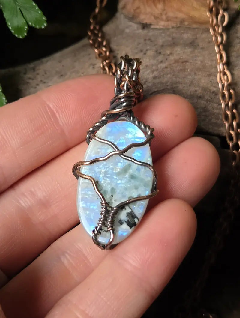 A close-up of a hand holding a copper-wrapped pendant necklace with an iridescent moonstone, with part of the copper chain visible against a dark natural background.