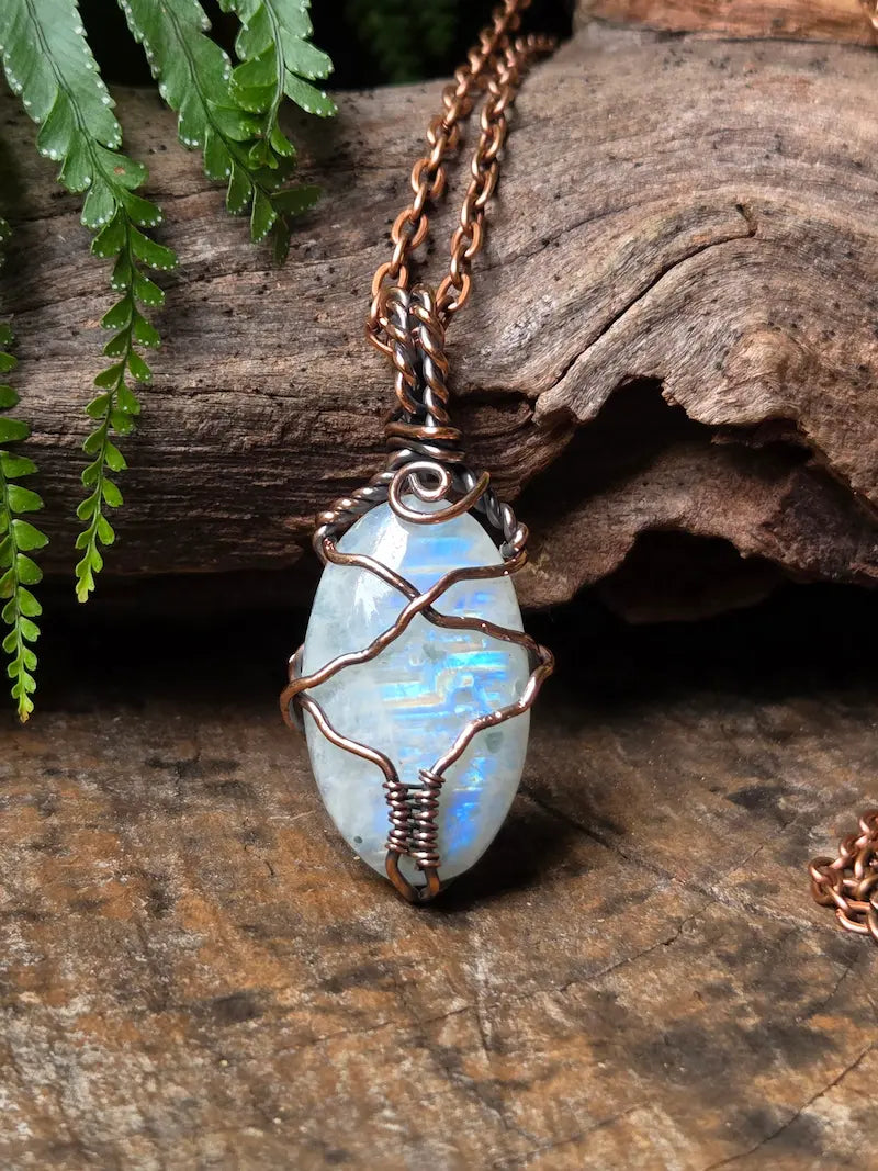 A handmade pendant necklace featuring an oval iridescent white gemstone wrapped in copper wire, hanging from a copper chain on a rustic wood background with green fern leaves.