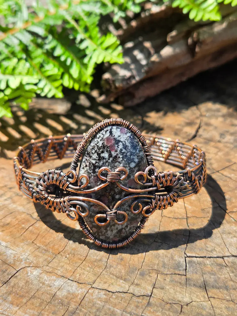 Copper wire wrapped bracelet with a Eudialyte stone, resting on a natural wooden surface with green ferns in the background 