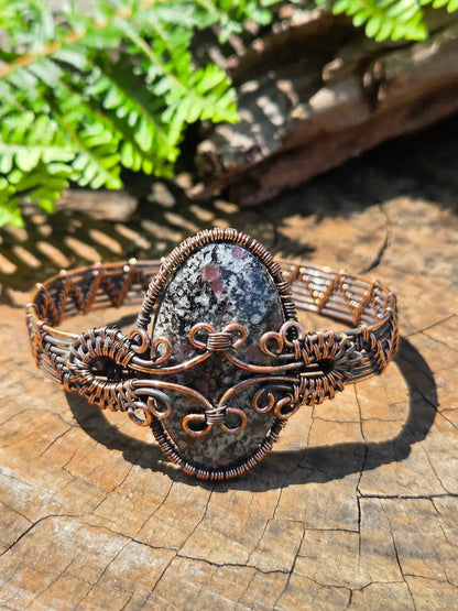 Copper wire wrapped bracelet with a Eudialyte stone, resting on a natural wooden surface with green ferns in the background 