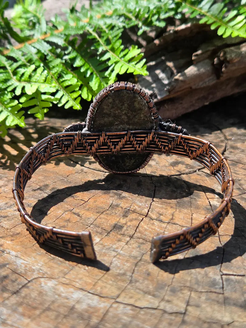 Copper wire wrapped bracelet with a Eudialyte stone, resting on a natural wooden surface with green ferns in the background 