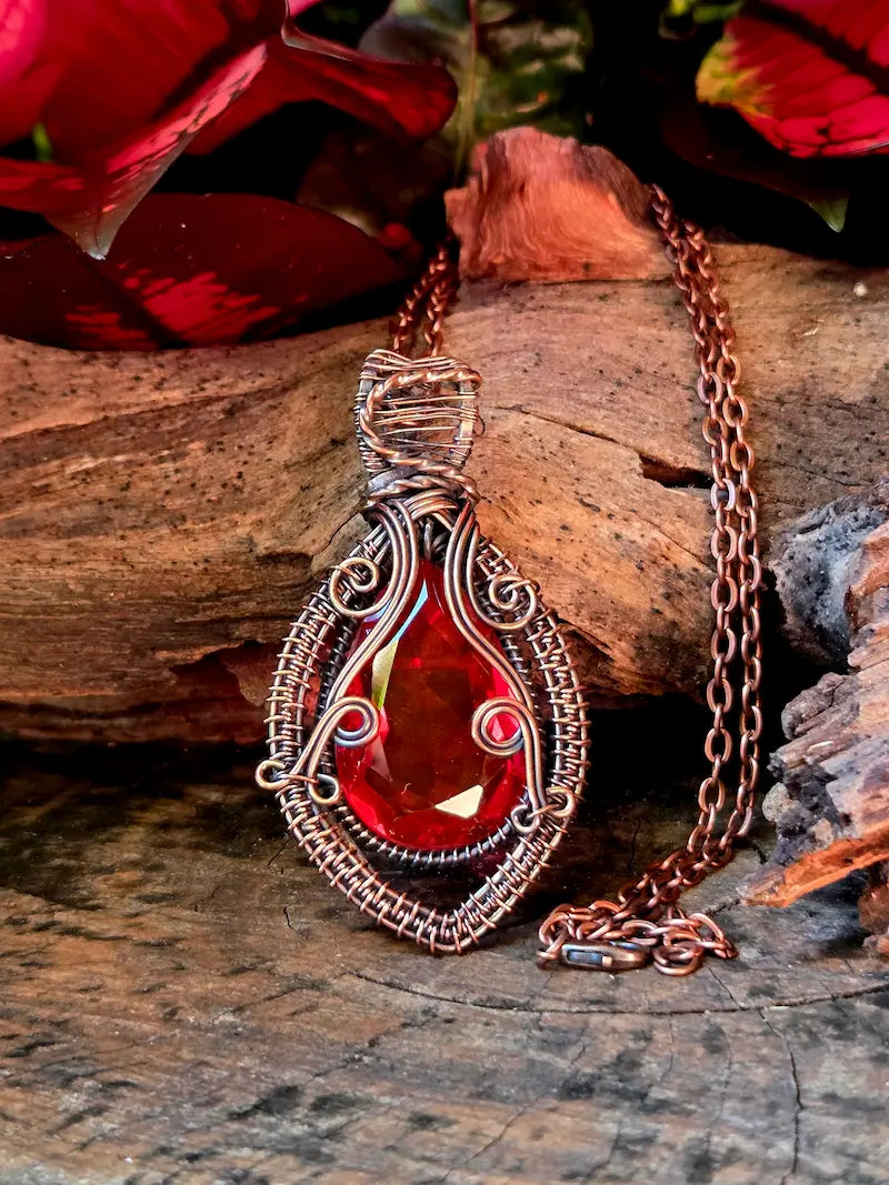 Red garnet copper pendant necklace on a wooden surface with red flowers in the background