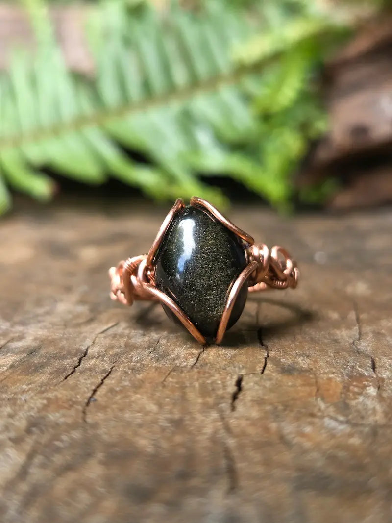 A handmade copper wire ring featuring a black Obsidian stone, resting on a textured wooden surface with a blurred green fern in the background.