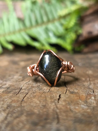 A handmade copper wire ring featuring a black Obsidian stone, resting on a textured wooden surface with a blurred green fern in the background.