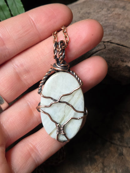 A handmade oval pendant featuring a pale white stone wrapped in copper wire, held in a person's hand against a dark rustic wood background.