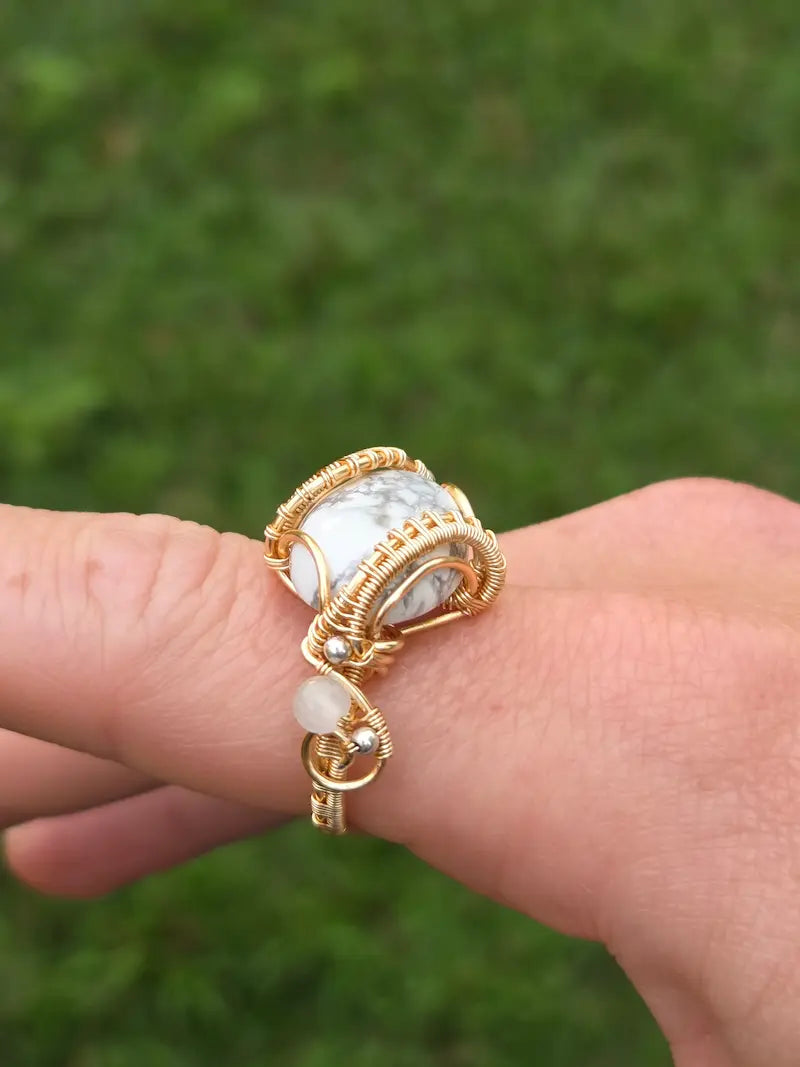 A copper wire-wrapped ring with a large white and gray marbled stone, worn on a person's finger against a blurred green outdoor background.